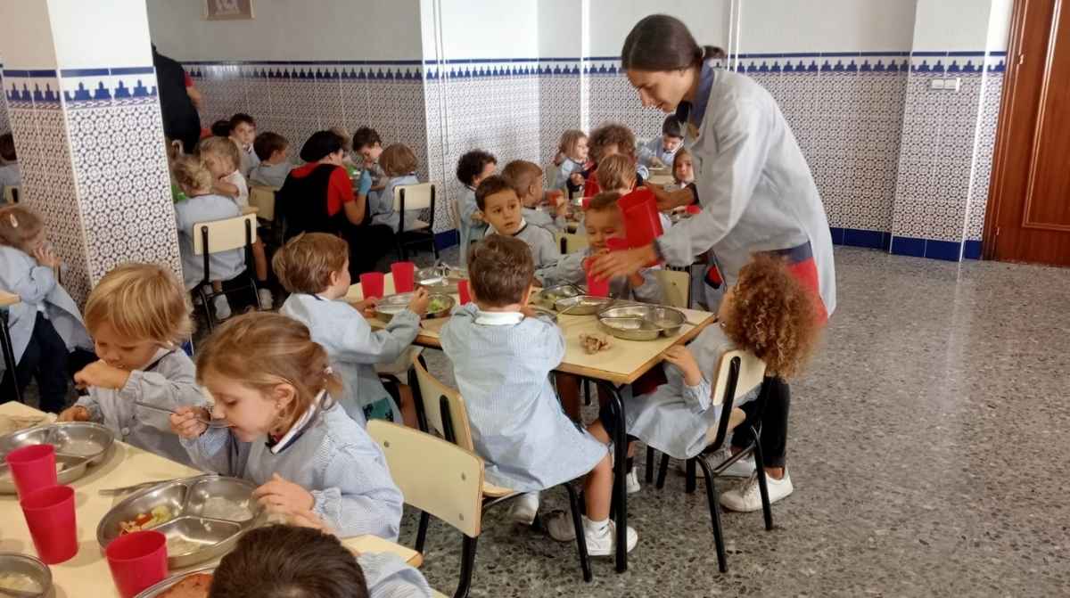 Comedor escolar con monitora atendiendo a niños durante la comida en un colegio