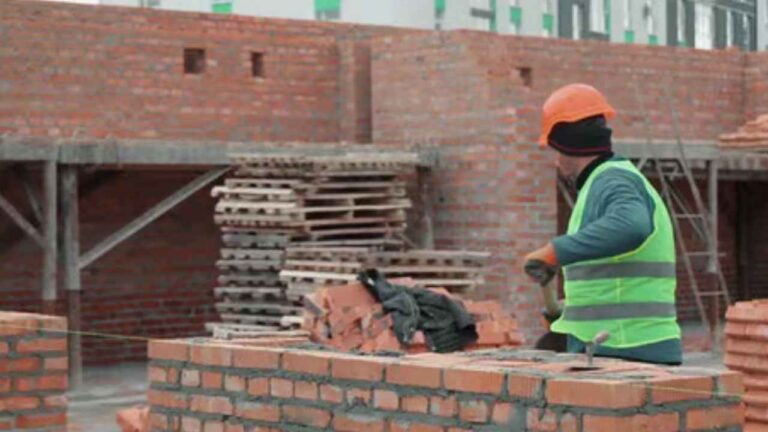 Albañil construyendo un muro de ladrillos en una obra mientras trabaja con casco y chaleco de seguridad.