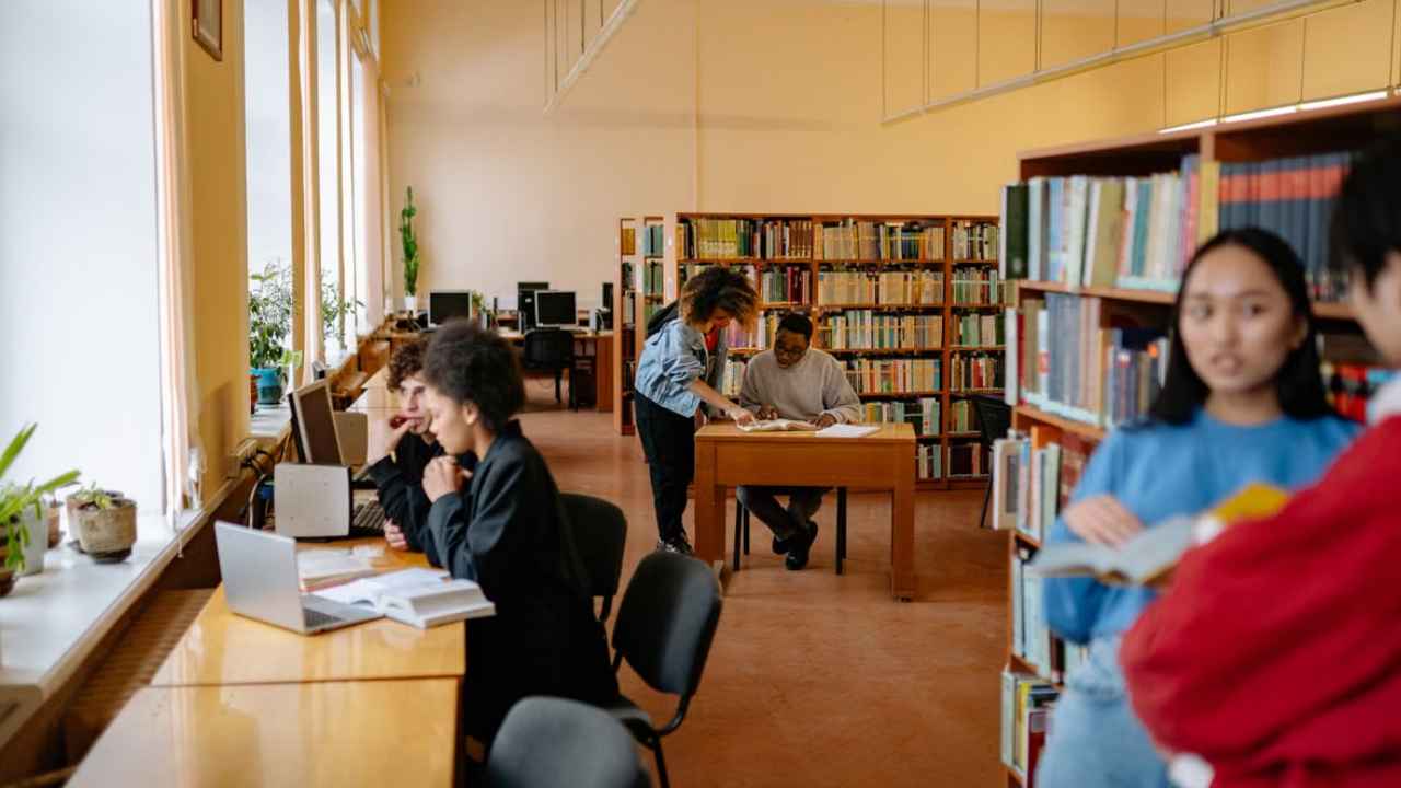 Personas estudiando entre mesas y estanterías de una biblioteca.