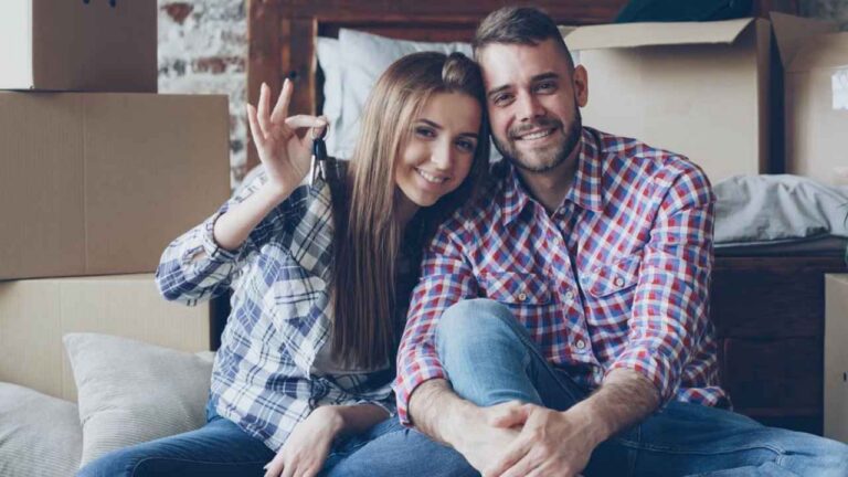 Una pareja sentada entre cajas de mudanza; ella muestra las llaves de una vivienda.