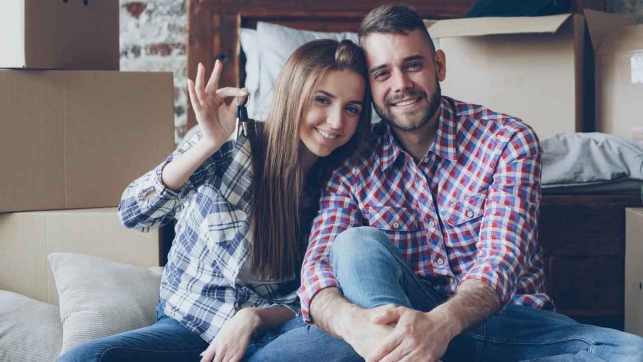 Una pareja sentada entre cajas de mudanza; ella muestra las llaves de una vivienda.