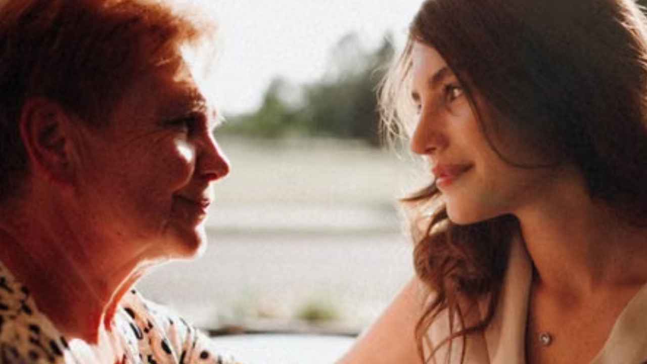 Una mujer mayor y otra mujer adulta se miran mientras se sujetan las manos junto a una ventana.