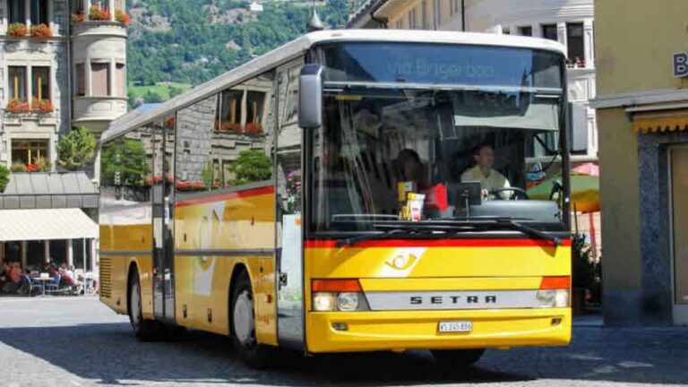 Autobús amarillo Setra en una plaza con edificios y montañas al fondo.
