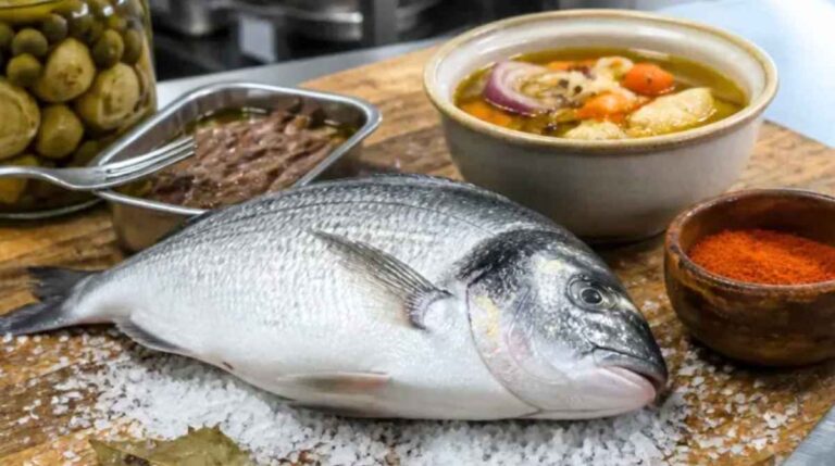 Pescado entero sobre sal gruesa, junto a tarros de conservas vegetales y otros alimentos en una cocina.