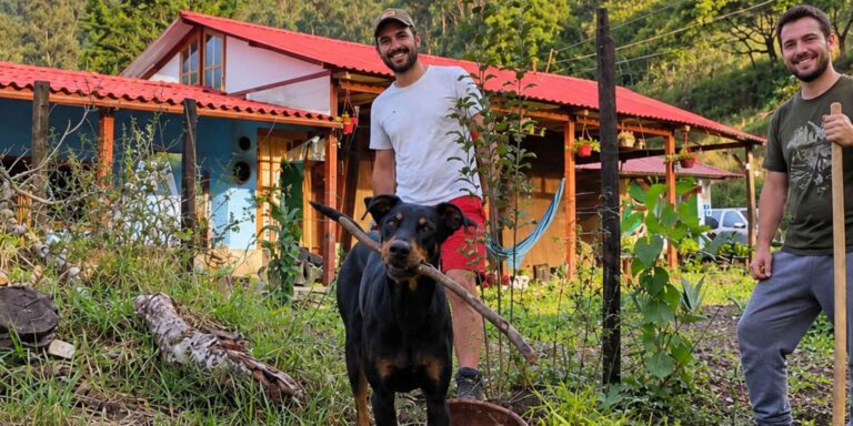 Dos hombres acompañados de un perro en una finca rural con jardín y vivienda, imagen ilustrativa del trabajo de mantenimiento de fincas. Copiar