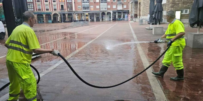 Dos operarios de limpieza viaria con uniformes amarillos limpiando con agua a presión el pavimento de una plaza. Copiar