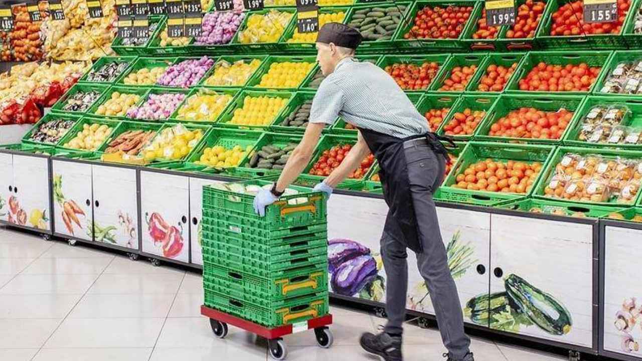 Trabajador de supermercado transportando cajas verdes de mercancía junto a la sección de frutas y verduras.