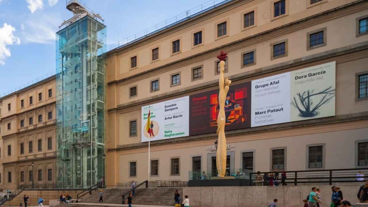 Fachada principal del Museo Reina Sofía de Madrid, con su ascensor de cristal y visitantes en la entrada.