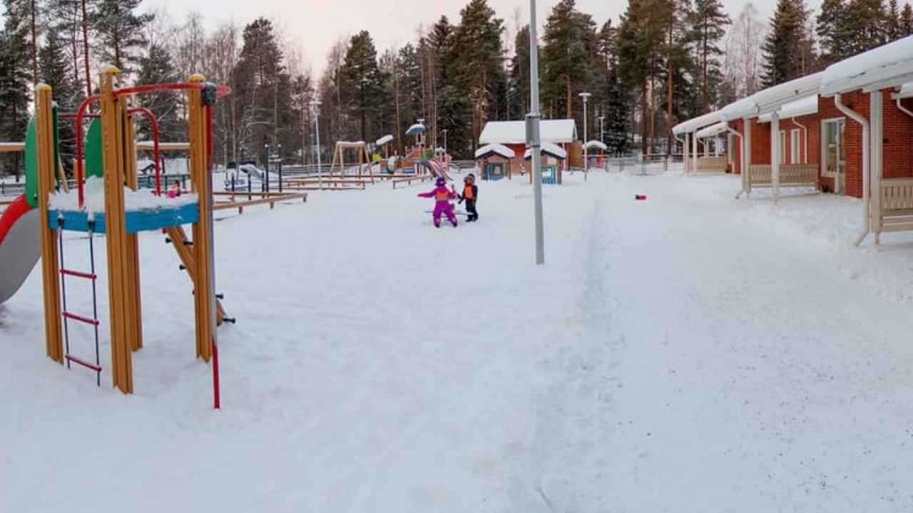 Patio de una escuela infantil cubierto de nieve, con tobogán, columpios y un edificio de ladrillo.