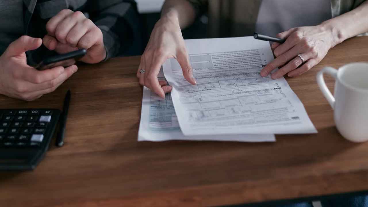 Dos personas revisan documentos junto a una calculadora y un teléfono sobre una mesa.