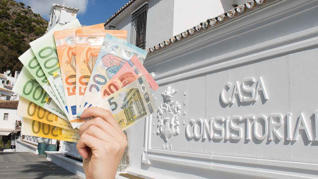Mano sosteniendo billetes de euro frente a la Casa Consistorial de Mijas, municipio que ofrece ayudas por discapacidad.