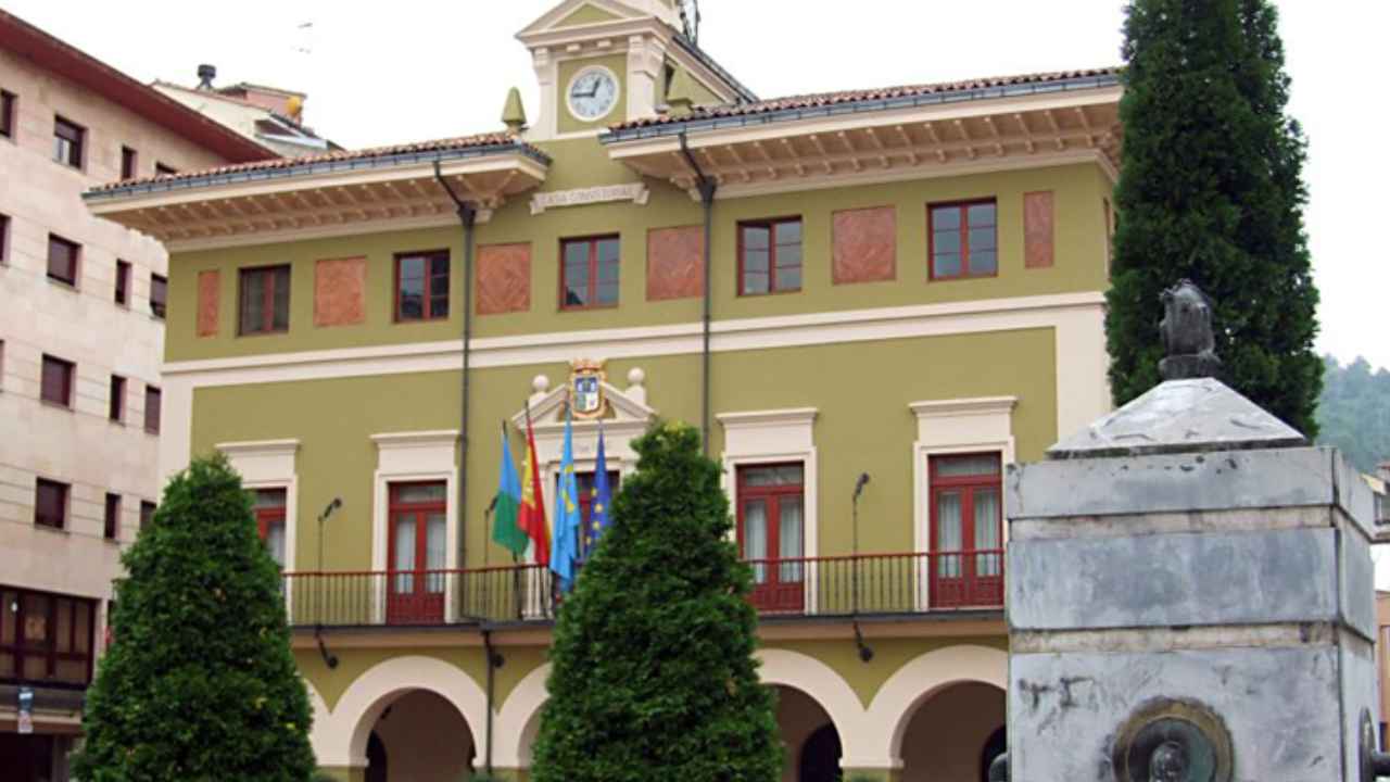 Fachada del Ayuntamiento de Langreo, en Asturias, con las banderas oficiales y una fuente en la plaza situada frente al edificio.