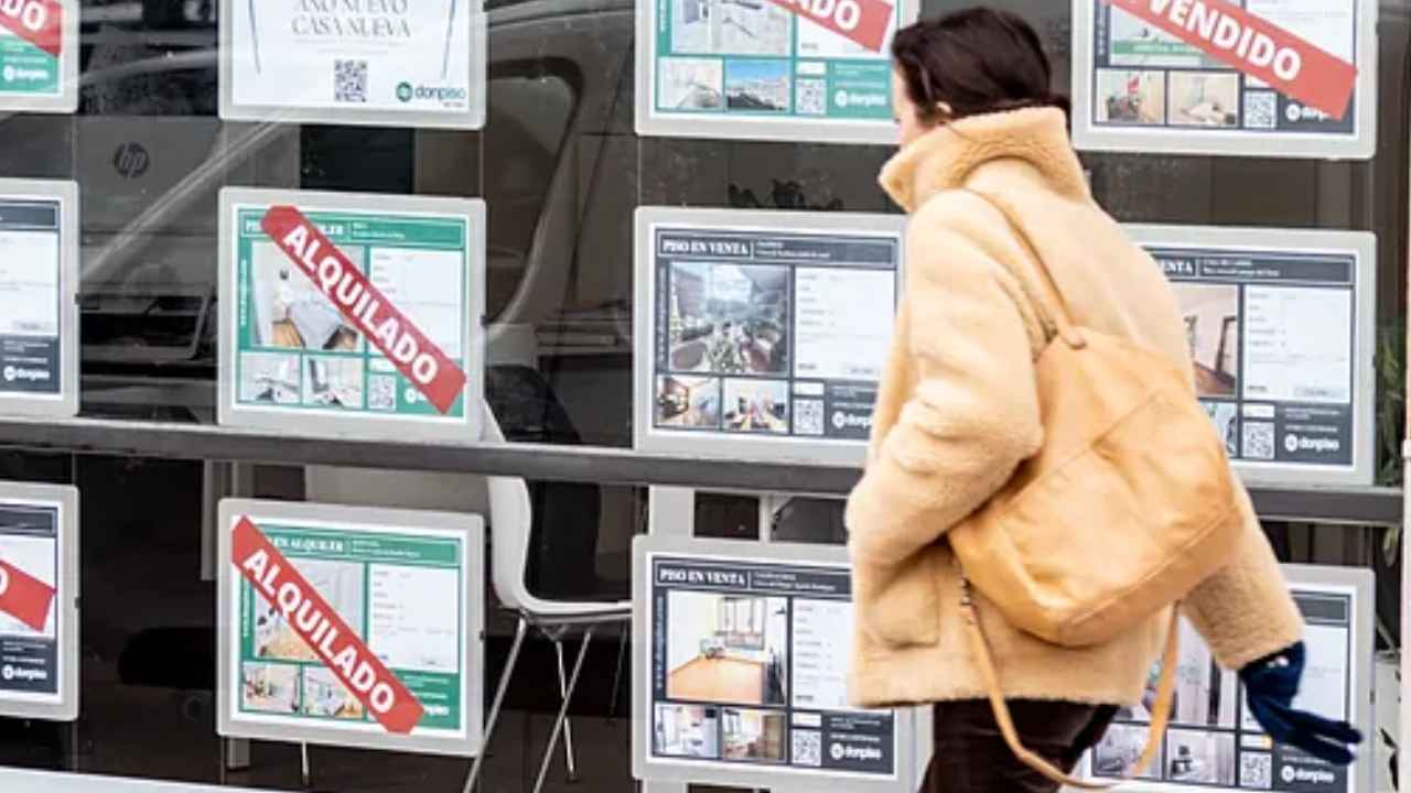 Mujer caminando frente a una inmobiliaria con anuncios de pisos alquilados y vendidos, reflejo de las dificultades de acceso a la vivienda.