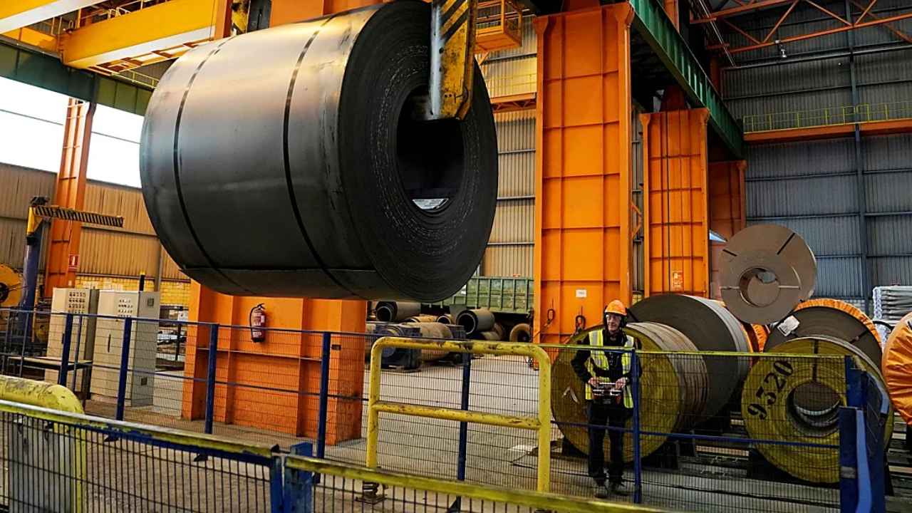 Trabajador en una nave industrial junto a grandes bobinas de acero y maquinaria de elevación.